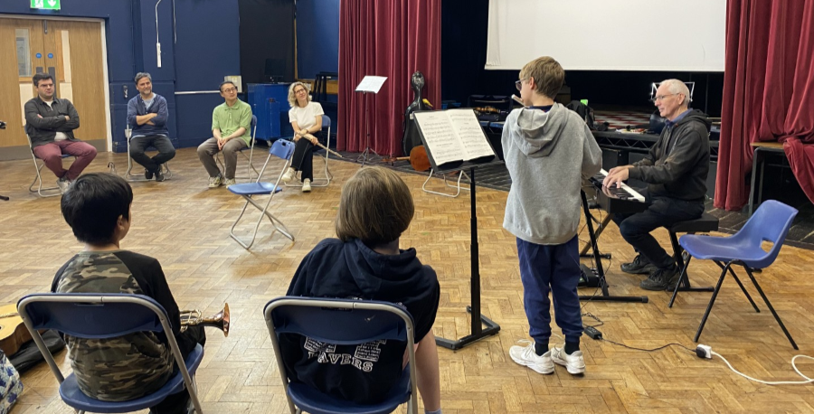 A boy performing in front of a music stand with a keyboard accompanist, as others watch in a school hall.